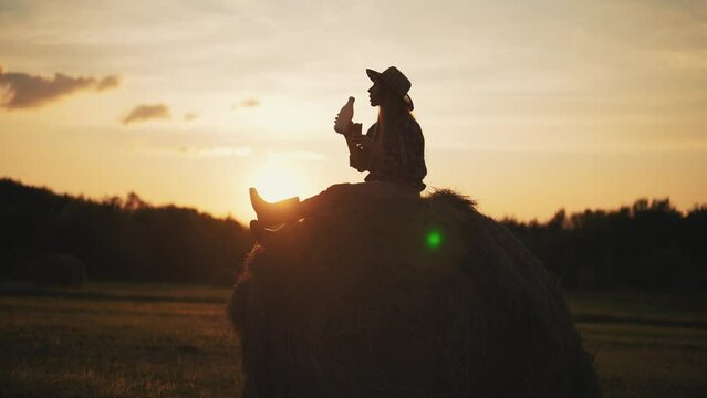 Silhouette of woman farm worker having dinner, drinking milk from bottle sitting on haystack on agricultural field at sunset. Daily routine of female farmer, farmland lifestyle, autumn harvesting.