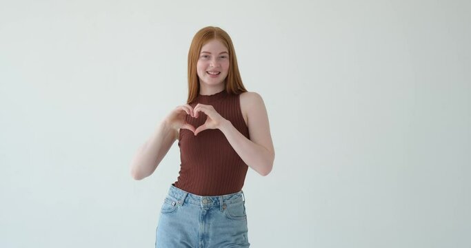 Red haired girl is portrayed showing a heart shape with her hands on a white background. With a warm and affectionate expression, she forms a heart with her fingers, symbolizing love and care.