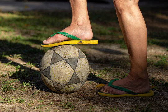 Pés de uma mulher dominando uma bola de futebol, com um dos pés em cima da bola em campo de grama desgastado.