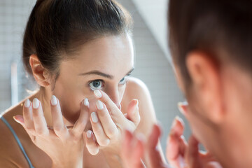 Mid adult woman applying contact lens in bathroom mirror.