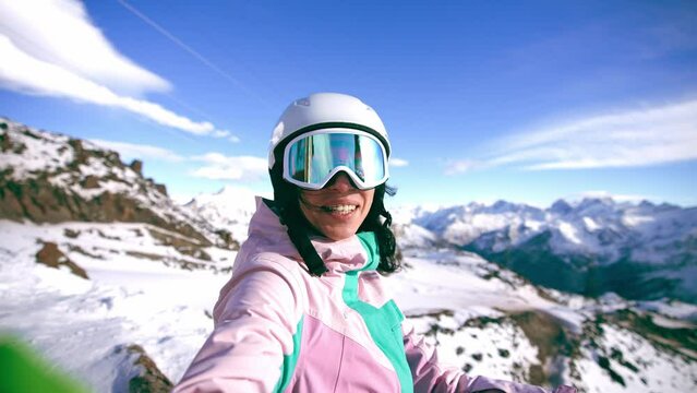 Smiling woman blogger vlogger standing on top mountain talking with camera recording video. Female skyer in protective clothing helmet goggles. Create content for blog, vlog. Winter sport resort.