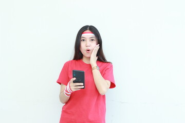 Naklejka premium portrait of beautiful asian woman wearing red outfit celebrating Indonesia independence day holding mobile phone with surprised expression
