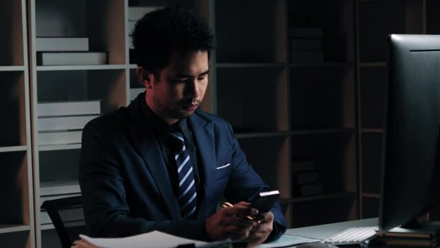 Tired young Asian businessman working overtime in the office room at night, trying to get things done before the deadline.