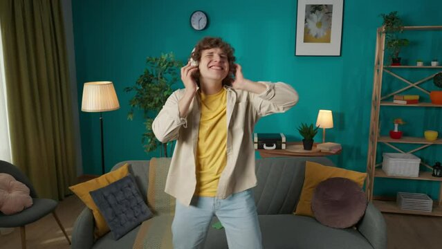 Man in casual clothing with headphones dancing happily in the living room, listening to music