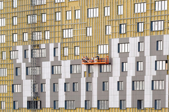 Workers team on construction cradle cladding and warming wall of monolithic house under construction. Man on suspended platform insulating wall facade of residential building..