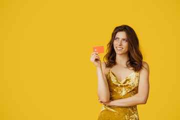 Cheerful beautiful woman in elegant dress holding credit card and dreaming about buying, yellow background, free space