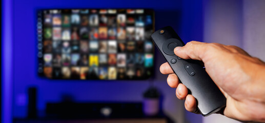 remote control in hand and tv with movie list on the screen in background. streaming media service, content on demand. banner with copy space - obrazy, fototapety, plakaty