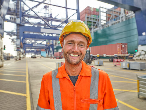 Smiling dock worker in reflective clothing and hard hat infront of container ship and cranes portrait