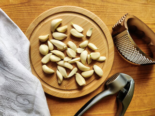 Peeled garlic cloves on chopping board, next to garlic choppers and gray linen towel on old wooden background. Concept of cooking and folk medicine for viral and colds. Top view. Copy space.