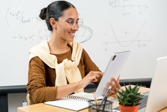 Teacher posing with tablet pc at elementary school. Young teacher of Latin appearance in cardigan glasses holds a tablet in her hand. Looks at the device, prints and prepares for the lesson