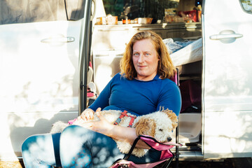 Portrait of middle-aged woman holding her cockapoo puppy pet while sitting near van camper car...