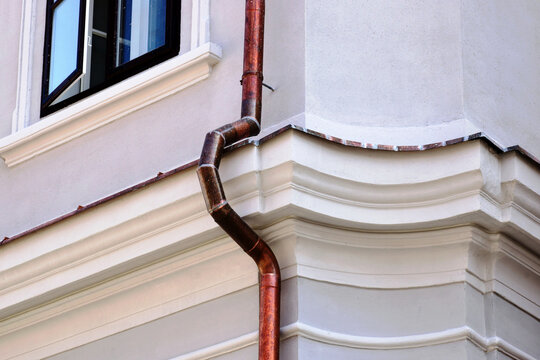 shiny new copper rain water leader, downspout or down pipe detail. lightning protection grounding rod. home renovation, construction concept. textured white stucco exterior wall and window background