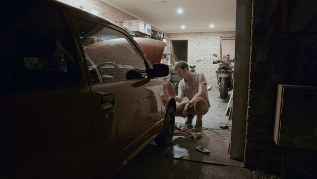 Young adult man dedicates time in his garage to polish and restore the headlights of his car, enhancing their clarity and appearance. Male is maintaining the overall condition of his vehicle's