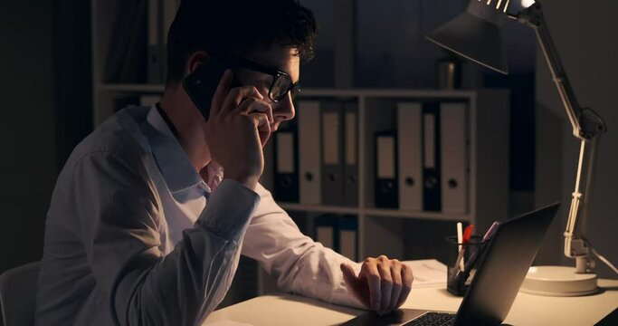 Dedicated businessman is engaged in a phone call while simultaneously working on laptop. The soft glow of the desk lamp illuminates his focused expression as he communicates with a sense of purpose.