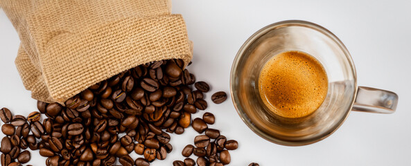Top view of hot black fresh coffee with foam and golden cream in glass cup with roasted arabica coffee beans in burlap sack bag on white background. Banner web.