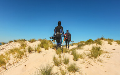 Two local Malagasy fishermen in neoprene wetsuit, with snorkel and simple harpoon, walking barefoot over wet sand beach, view from behind