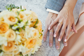 Hands of the bride and groom with wedding rings and a wedding bouquet in the foreground.