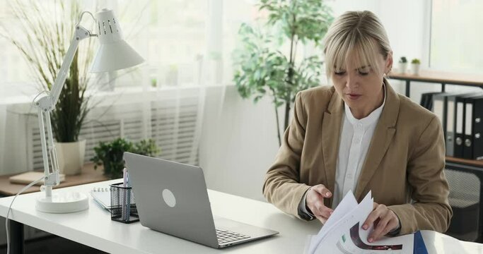 Self-assured woman in an office setting, proficiently managing both a laptop and papers. With a poised and focused attitude, she seamlessly transitions between tasks.