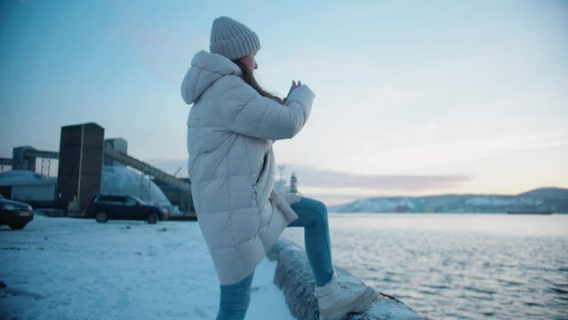 A young caucasian girl in a winter jacket, long hair and a white hat walks and puts her foot in blue jeans on the ledge and takes pictures of the winter sea and hills on smartphone
