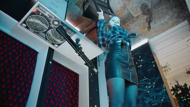 A caucasian slim young girl in a black leather skirt, white sneakers, tights and a dark shirt stands in the house on the contrary and holds on to a chair attached to the brown ceiling