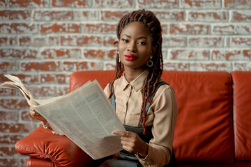 Attractive black woman sitting on the orange leather sofa holding a newspaper in her hands and looking at the camera. Young citizen checking on the local news. Entrepreneur, content writer, sub-editor - obrazy, fototapety, plakaty