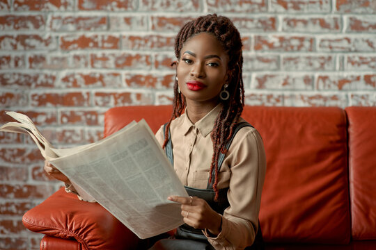 Attractive black woman sitting on the orange leather sofa holding a newspaper in her hands and looking at the camera. Young citizen checking on the local news. Entrepreneur, content writer, sub-editor