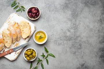 Sliced fresh ciabatta on cutting kitchen board, green and brown olives, olive oil with rosemary, olive tree branches on gray concrete stone rustic background from above copy space - 633125195
