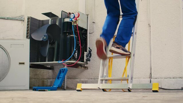 Meticulous mechanic in professional uniform with technical equipment climbing folding ladder to do maintenance on rooftop hvac system. Professional worker commissioned to check up on air conditioner
