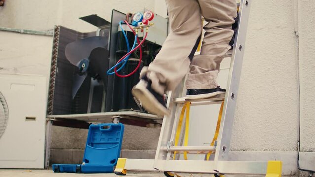 Ground level shot of capable electrician holding hand drill climbing folding ladder to do maintenance on rooftop hvac system. Seasoned wireman commissioned to checkup on condenser