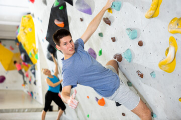 Young man making thumb up gesture and looking at camera while climbing on rock-climbing wall during training in bouldering gym. - obrazy, fototapety, plakaty