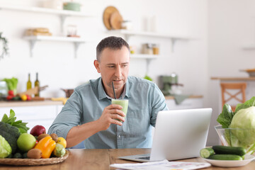 Mature man with glass of fresh vegetable smoothie using laptop in kitchen