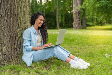 Obraz premium Happy Middle Eastern female student using laptop computer outdoors in the park. Girl learning tasks for preparing for exams. Woman with curly dark hair studying material for university