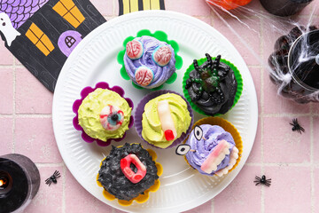 Plate with delicious Halloween cupcakes and candies on pink tile background, closeup