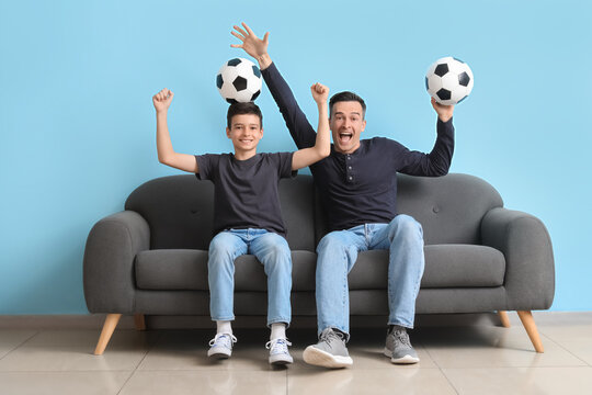 Happy little boy with his dad watching football game on sofa near blue wall