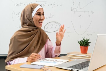 Young Eastern female teacher in hijab greeting students by waving hand during online lesson while sitting by desk in front of laptop. Smiling Islamic tutor in classroom. Education learning concept - obrazy, fototapety, plakaty