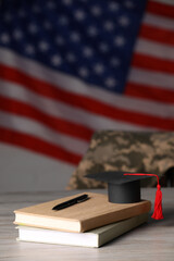 Notebooks, mortarboard and pen on wooden table against flag of USA. Military education