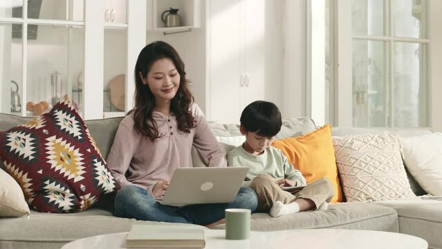 young asian woman working at home with son playing computer game by her side
