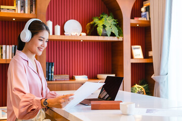 Happy asian businesswoman  looking at paper while watching webinar or doing video chat by webcam.Asian female entrepreneur working remotely from home office using modern computer.