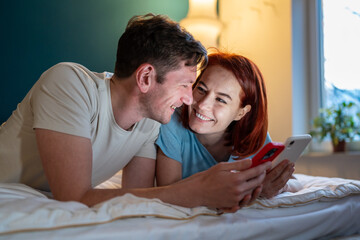 Couple in love smiles, lying on bed with smartphones. Happy woman and man lying side by side with mobile phones and looking into each other's eyes. Expression of joy fun love on faces of guy and girl.