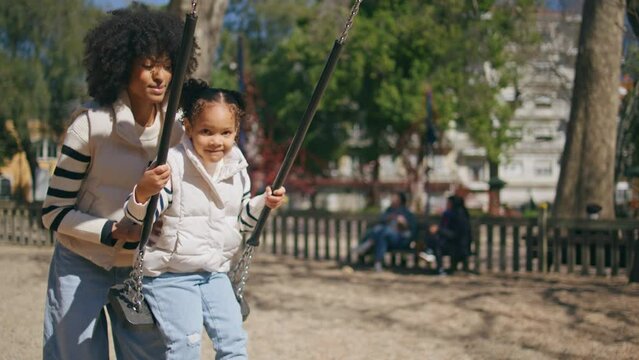 Mother pushing swing daughter playing on playground. Cute african girl swinging.