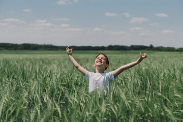 Naklejka na ściany i meble Portrait of a smiling boy in a white T-shirt and playing in a green barley field. Happy child boy laughing and playing in the summer day. Kid exploring nature. Summer activity for inquisitive children
