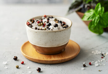 Sea salt with mix of peppercorns in small ceramic bowl over small wooden plate, aromatic raw herbs on concrete background