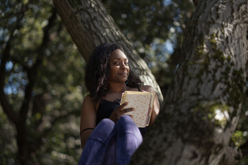 A person relaxes, enjoying a moment alone in a tree to sit and write in their notebook. They have their hair down and are smiling, express their joy in a reflective, introspective moment.