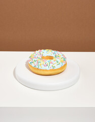 A deliciously appetizing doughnut with fresh glaze and colorful sprinkles lies on a white round stand.