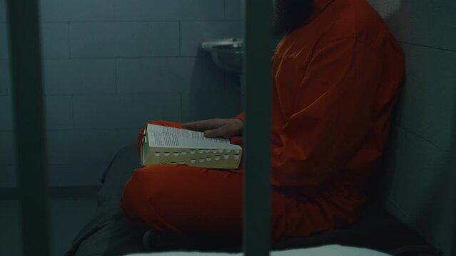 Male prisoner in orange uniform sits on bed, reads Bible in prison cell. Criminal, inmate serves imprisonment term for crime in jail. Detention center or correctional facility. Faith in God concept.