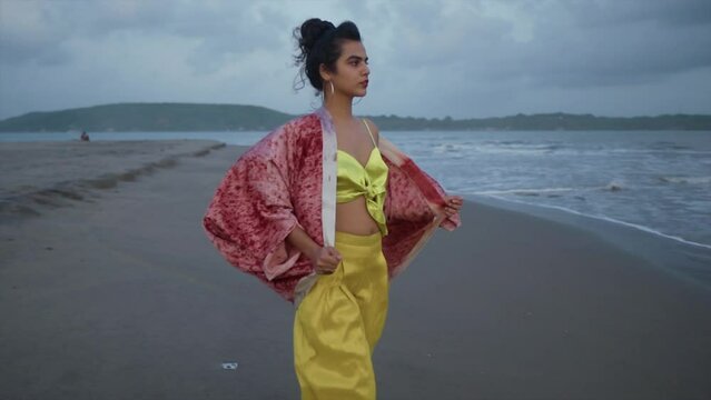 Cinematic footage of an Indian fashion model wearing a yellow outfit and a red silk jacket on a sandy beach at the ocean on a cloudy day in Goa India, Slow Motion, Slomo
