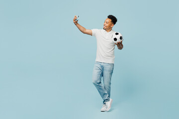 Full body young man fan wear t-shirt cheer up support football sport team hold in hand soccer ball watch tv live stream doing selfie shot on mobile cell phone isolated on plain blue color background.