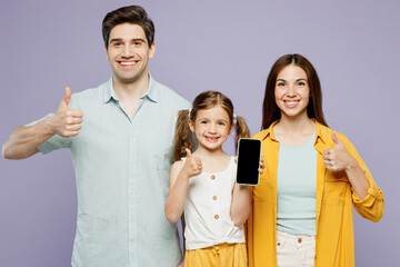 Young parents mom dad with child kid girl 6 years old wearing blue yellow casual clothes hold use blank screen mobile cell phone show thumb up isolated on plain purple background. Family day concept.