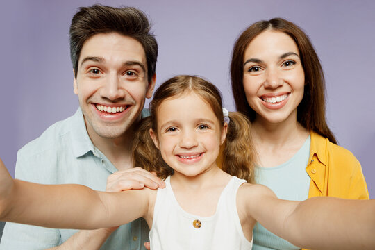 Close up young cool parents mom dad with child kid daughter girl 6 year old wear blue yellow casual clothes do selfie shot pov mobile cell phone isolated on plain purple background Family day concept.