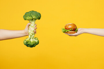 Close up cropped female hold in hand broccoli vegetable against burger fast food isolated on plain yellow color background studio. Healthy lifestyle motivation concept. Copy space advertising mock up.